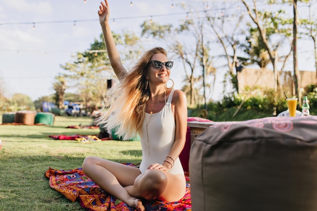 Foto gratuita mujer ligeramente bronceada en traje de baño sentada en el césped y agitando la mano. retrato al aire libre de una hermosa joven con cabello rubio divirtiéndose en el parque.