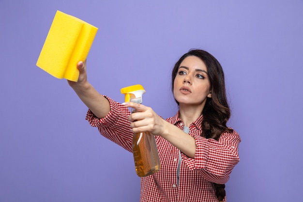 Foto gratuita mujer limpiador bastante caucásico confidente que sostiene el limpiador en aerosol y una esponja mirando al lado