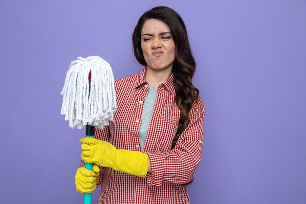 Foto gratuita mujer limpiadora bastante caucásica disgustada con guantes de goma sosteniendo y mirando la fregona