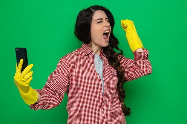 Mujer limpiadora bastante caucásica emocionada con guantes de goma sosteniendo el teléfono y levantando el puño de pie con los ojos cerrados