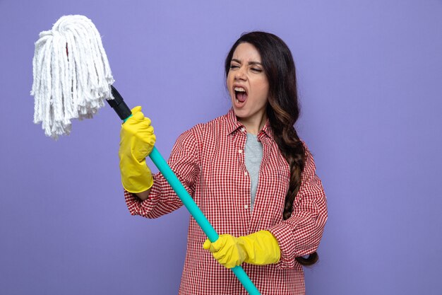 Foto gratuita mujer limpiadora bastante caucásica molesta con guantes de goma sosteniendo y mirando la fregona