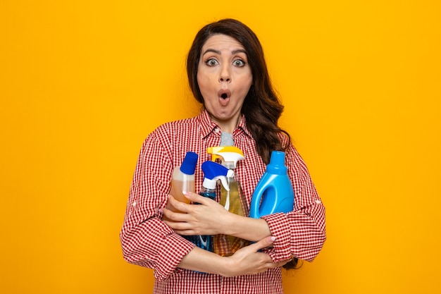 Mujer limpiadora bastante caucásica sorprendida que sostiene los aerosoles y líquidos de limpieza