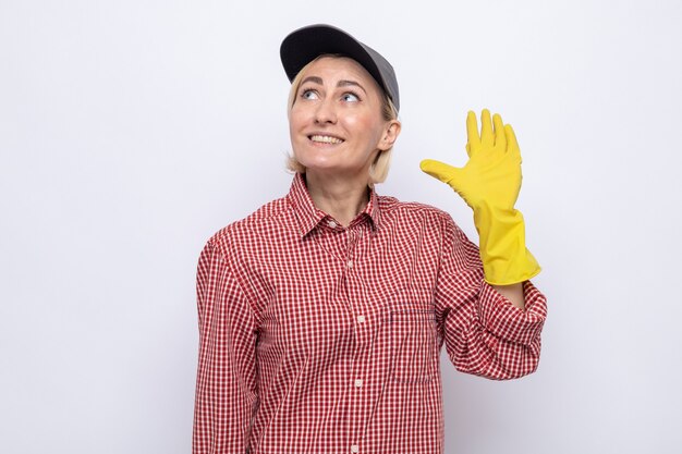 Mujer de la limpieza en camisa a cuadros y gorra con guantes de goma mirando hacia arriba sonriendo alegremente saludando con la mano