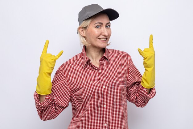 Mujer de la limpieza en camisa a cuadros y gorra con guantes de goma mirando a la cámara feliz y alegre mostrando el símbolo de la roca sobre fondo blanco.
