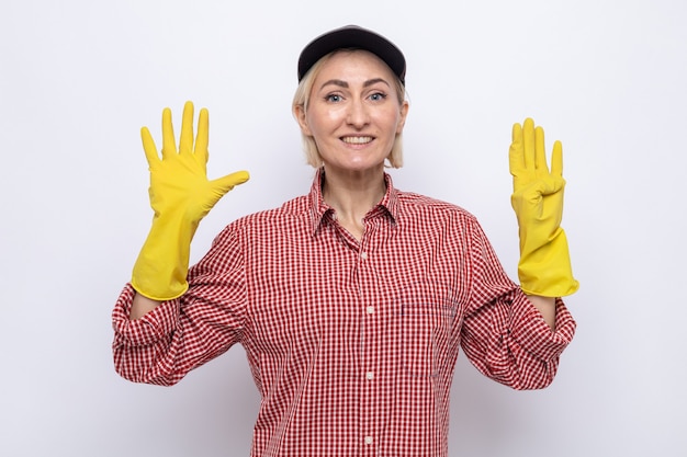 Mujer de la limpieza en camisa a cuadros y gorra con guantes de goma mirando sonriendo alegremente mostrando el número nueve con los dedos