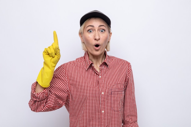 Mujer de la limpieza en camisa a cuadros y gorra con guantes de goma mirando sorprendido apuntando con el dedo índice hacia arriba
