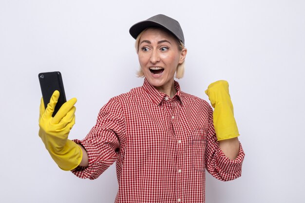 Mujer de la limpieza en camisa a cuadros y gorra con guantes de goma mirando su teléfono móvil feliz y emocionado puño apretado