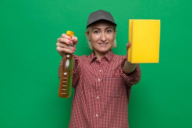 Foto gratuita mujer de la limpieza en camisa a cuadros y gorra sosteniendo una esponja y spray de limpieza mirando sonriente seguro listo para la limpieza