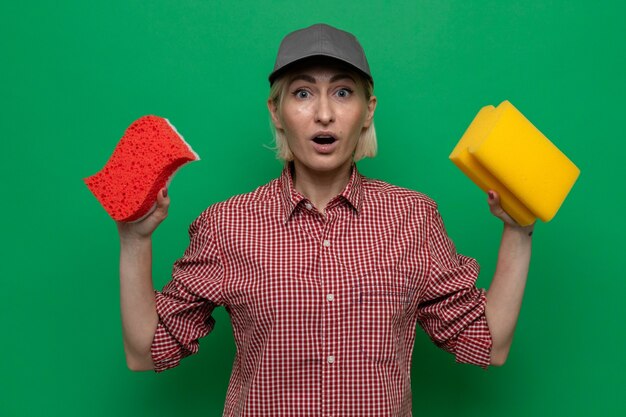 Mujer de la limpieza en camisa a cuadros y gorra sosteniendo esponjas mirando a la cámara asombrado y sorprendido de pie sobre fondo verde