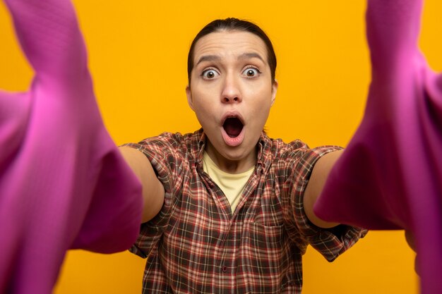 Foto gratuita mujer de limpieza joven en camisa a cuadros y gorra en guantes de goma tomando selfie asombrado y sorprendido de pie en rosa