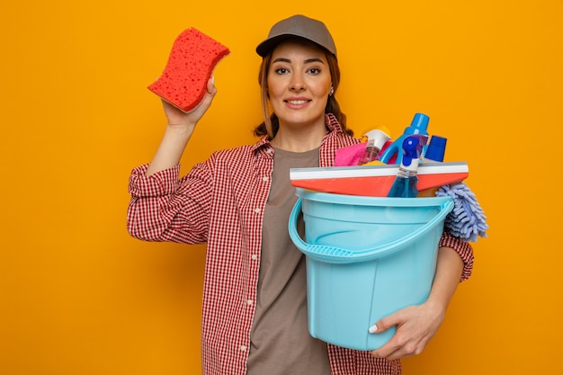 Mujer de limpieza joven en camisa a cuadros y gorra sosteniendo un balde con herramientas de limpieza mostrando esponja sonriendo confiada mirando a la cámara de pie sobre fondo naranja