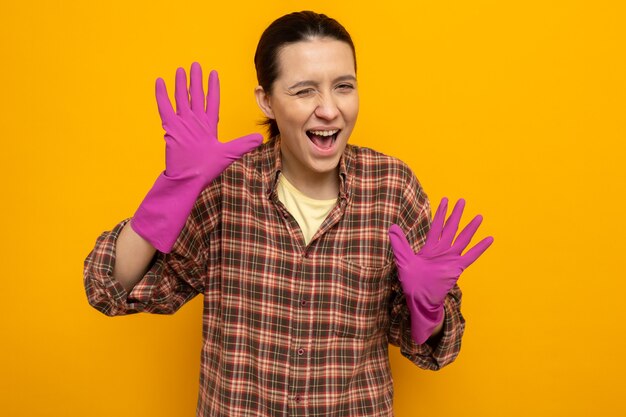 Mujer de limpieza joven en camisa a cuadros en guantes de goma mirando al frente sonriendo y guiñando un ojo mostrando palmas de pie sobre la pared naranja