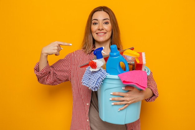 Mujer de limpieza joven feliz en camisa a cuadros sosteniendo un balde con herramientas de limpieza mirando a la cámara sonriendo alegremente apuntando con el dedo índice a sí misma de pie sobre fondo naranja