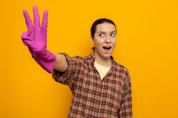 Mujer de limpieza joven feliz y emocionada en ropa casual en guantes de goma que muestra el número tres con los dedos de pie sobre la pared naranja