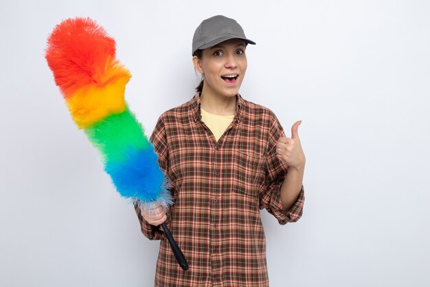 Mujer de limpieza joven feliz en ropa casual y gorra sosteniendo plumero colorido sonriendo alegremente mostrando los pulgares para arriba de pie en blanco