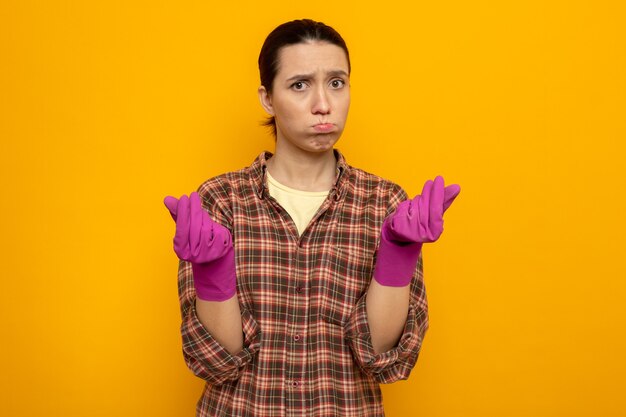Mujer de limpieza joven en ropa casual con guantes de goma disgustado soplando mejillas frotándose los dedos de pie sobre la pared naranja