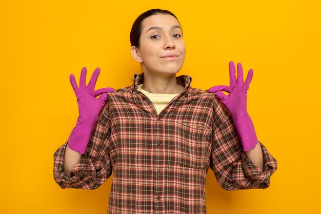 Mujer de limpieza joven en ropa casual en guantes de goma con expresión de confianza tocando su camisa de pie sobre la pared naranja