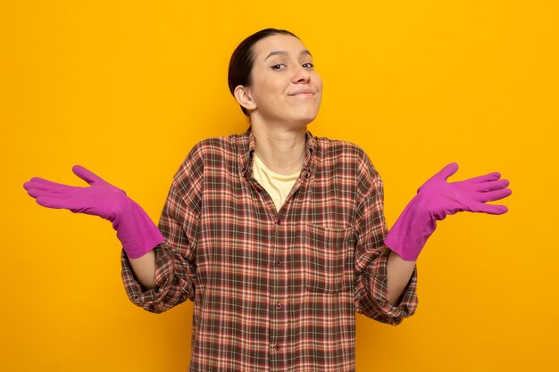 Mujer de limpieza joven en ropa casual en guantes de goma sonriendo extendiendo los brazos hacia los lados de pie en naranja