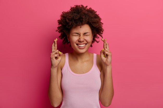 Foto gratuita la mujer linda y alegre y complacida se para con los dedos cruzados, quiere que los sueños se hagan realidad, cree en la buena suerte, usa un chaleco informal, pide un deseo, espera una respuesta positiva o buenas noticias, se mantiene esperanzada en el interior.