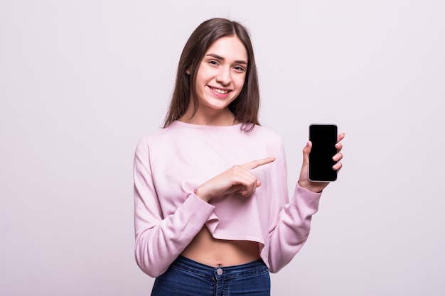 Mujer linda alegre que señala el dedo en la pantalla del teléfono inteligente aislada en un fondo blanco.
