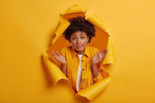 Una mujer linda incierta con peinado afro extiende las palmas de las manos, tiene una expresión confusa y despistada que toma una decisión, se encoge de hombros y no tiene idea de poses en un fondo de agujero de papel amarillo.