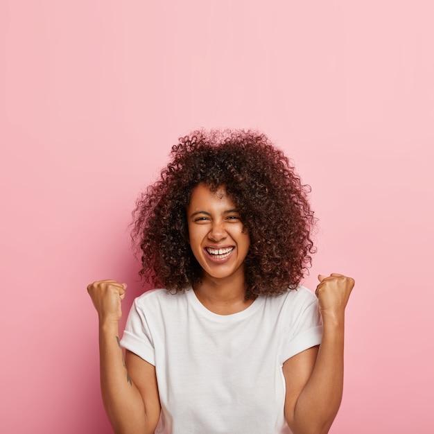 La mujer linda joven alegre y emocionada levanta los puños cerrados, se para sin maquillaje contra la pared rosada, tiene el pelo rizado y tupido, celebra la victoria y el triunfo, viste una camiseta blanca todos los días. ¡Oh si!