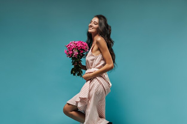 Mujer linda moderna con cabello ondulado en vestido elegante de verano a cuadros con ramo rosa y sonriendo sobre fondo azul aislado