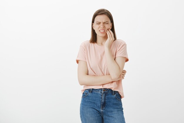 Mujer linda molesta que hace muecas de dolor y toca la mejilla, quejándose de dolor de muelas, necesita dentista