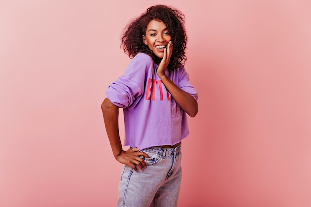 Mujer linda optimista con cabello ondulado que expresa emociones positivas. Filmación en interiores de la alegre dama negra divirtiéndose en rosa.