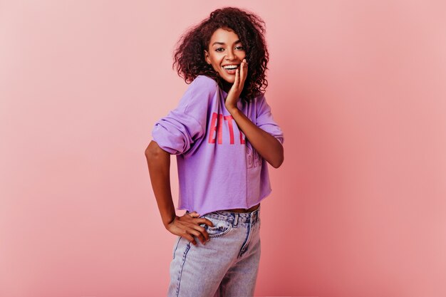 Mujer linda optimista con cabello ondulado que expresa emociones positivas. Filmación en interiores de la alegre dama negra divirtiéndose en rosa.