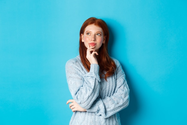 Mujer linda pensativa con el pelo rojo, mirando el logo de la esquina superior izquierda y pensando, imaginando algo, de pie sobre fondo azul.