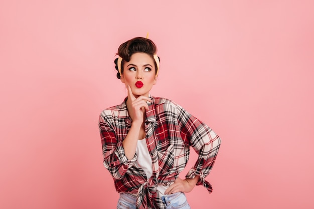 Mujer linda pensativa con pie de maquillaje brillante sobre fondo rosa. Disparo de estudio de chica pinup complacida en camisa a cuadros.