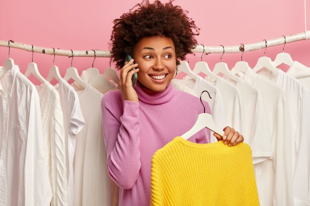 La mujer llena de alegría emocional hace una llamada telefónica, se para cerca del estante wardobe lleno de ropa blanca, sostiene un suéter amarillo de invierno de punto, disfruta del día de compras en el centro comercial de moda.