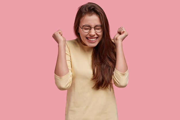 Mujer llena de alegría levanta los puños cerrados en señal de alegría, disfruta del éxito y el triunfo, usa anteojos y atuendo informal, modelos sobre espacio rosa