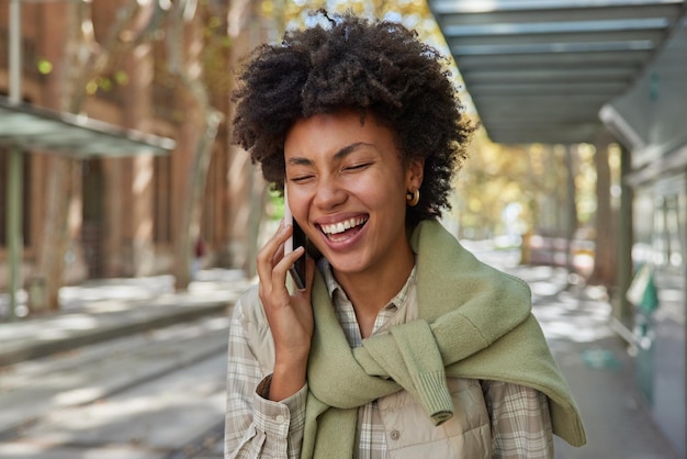 La mujer llena de alegría se ríe alegremente tiene una conversación telefónica vestida con ropa informal sonríe con dientes disfruta de una comunicación positiva pasa el tiempo libre al aire libre camina por la calle La chica habla por celular