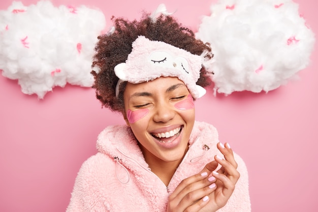 Mujer llena de alegría ríe feliz sonríe ampliamente cierra los ojos se despierta de buen humor usa máscara de dormir pijama cálido cierra los ojos de satisfacción aplica almohadillas de belleza para reducir las arrugas debajo de los ojos