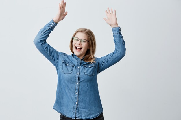 Una mujer llena de alegría usa una camisa de mezclilla, gafas elegantes, gestos de emoción, exclama en voz alta de asombro, se alegra de encontrarse con un viejo amigo y levanta los brazos. La mujer rubia disfruta el éxito en el trabajo.