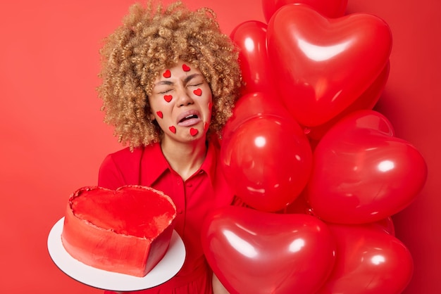 Foto gratuita una mujer llorando molesta con el pelo rizado se siente abatida sostiene un pastel dulce en forma de corazón y un montón de globos inflados ha estropeado el estado de ánimo durante el día de san valentín aislado sobre fondo rojo concepto de celebración