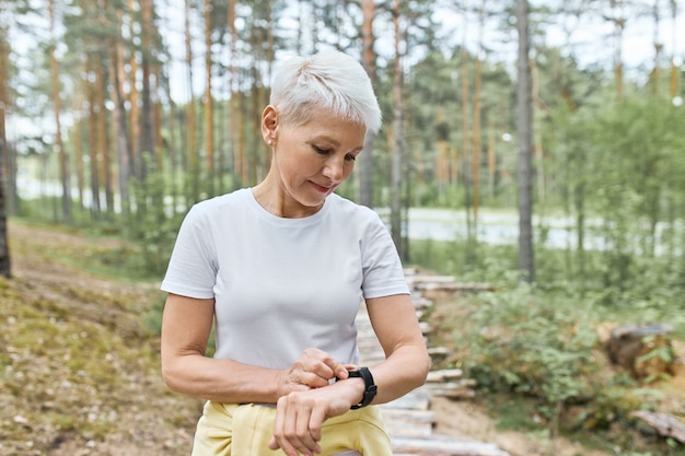 Mujer madura activa con cabello corto y rubio posando al aire libre, preparándose para hacer ejercicio, configurar el reloj inteligente, rastrear la frecuencia cardíaca y el pulso.