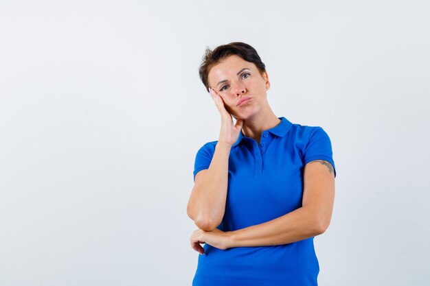 Mujer madura apoyándose en la palma de la mejilla en camiseta azul y mirando pensativo, vista frontal.