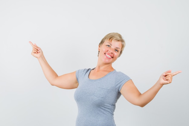 Mujer madura apuntando hacia afuera en camiseta gris y mirando alegre.