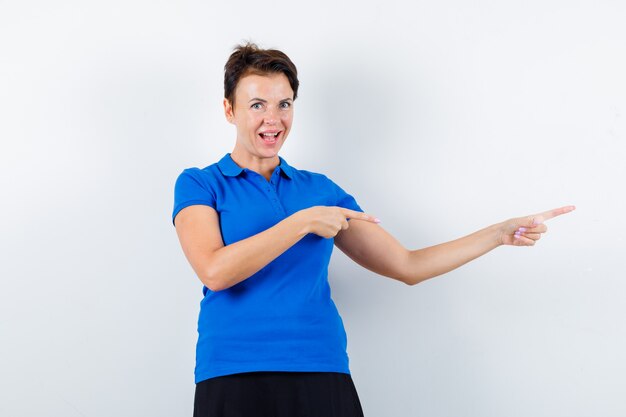 Mujer madura apuntando a la derecha en camiseta azul y mirando confiado, vista frontal.