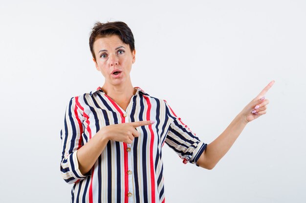 Mujer madura apuntando hacia la derecha con los dedos índices en camisa a rayas y mirando sorprendido, vista frontal.