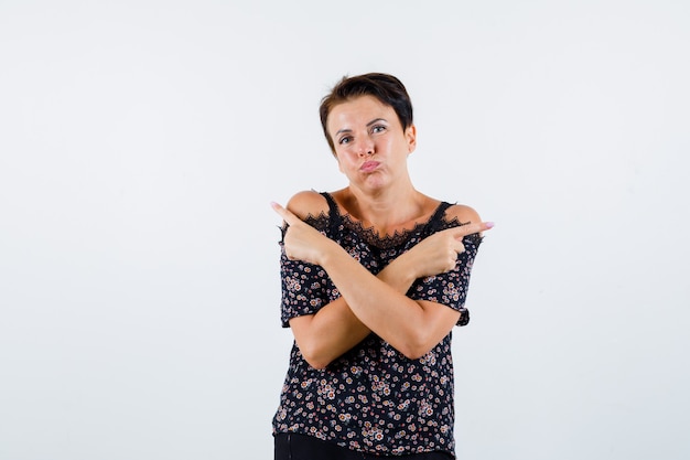 Mujer madura apuntando en direcciones opuestas con los dedos índices, hinchando las mejillas con blusa floral, falda negra y aspecto serio. vista frontal.