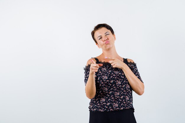 Mujer madura apuntando hacia la izquierda con el dedo índice, enviando besos en blusa floral y falda negra y luciendo alegre. vista frontal.