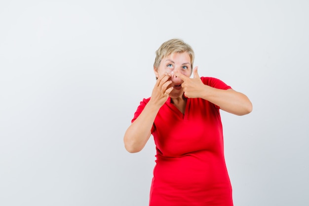 Mujer madura apuntando a su párpado tirado por el dedo en camiseta roja