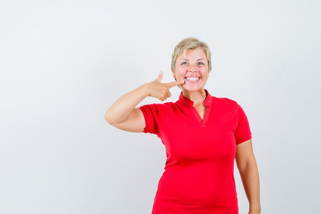 Mujer madura apuntando a sus dientes en camiseta roja y mirando contenta.