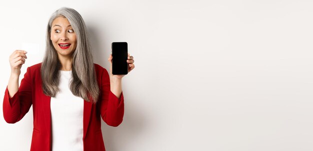 Mujer madura asiática alegre que muestra la pantalla del teléfono inteligente en blanco y el concepto de tarjeta de crédito de comercio electrónico