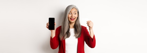 Foto gratuita mujer madura asiática alegre que muestra la pantalla del teléfono inteligente en blanco y el concepto de tarjeta de crédito de comercio electrónico