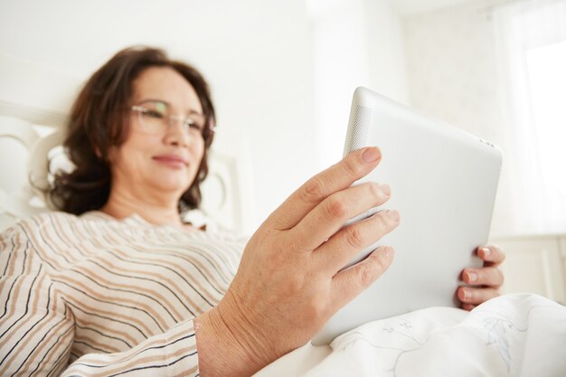 Mujer madura atenta y alegre con un tablet pc acostada en su cama en un dormitorio